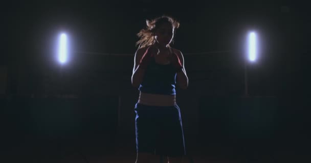 En regardant dans la caméra, une belle boxeuse frappe sur un fond sombre avec une lumière rétro-éclairée. Steadicam shot — Video