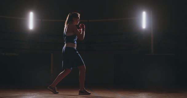 Una hermosa boxeadora se entrena en un gimnasio oscuro y hace ejercicios de golpes en cámara lenta. Vista lateral del movimiento de la cámara. Steadicam tiro — Vídeos de Stock