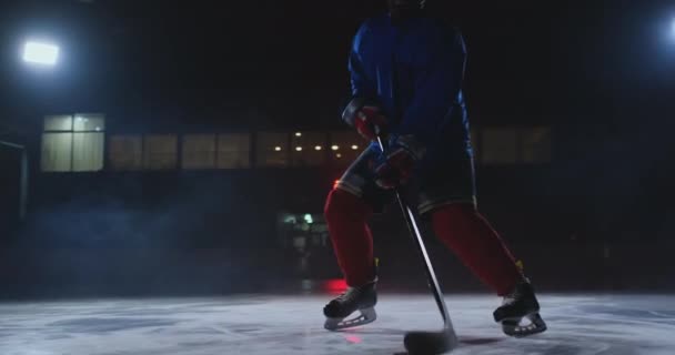 Jugador de hockey masculino con un disco en la arena de hielo muestra goteo moviéndose directamente en la cámara y mirando directamente a la cámara contra un fondo oscuro en el humo — Vídeo de stock