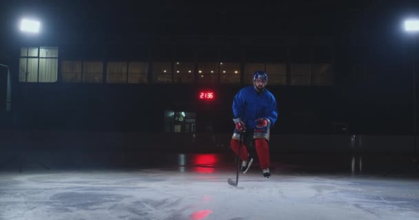 Hokeista człowiek w mundurze na lodzie pozostawia z kijem w ręku z ciemności i patrzy prosto w kamerę — Wideo stockowe