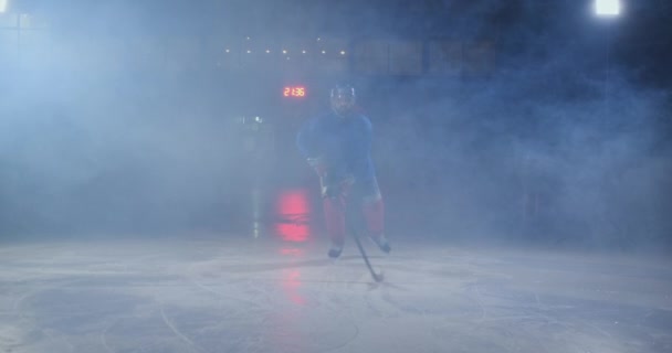 Человек хоккеист с шайбой на льду в хоккейной форме листья с клюшкой в руках из темноты и смотрит прямо в камеру — стоковое видео