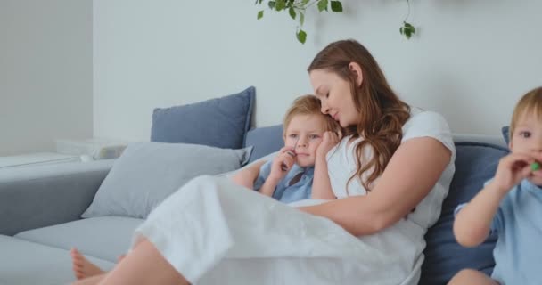 Maman étreint ses deux petits fils sur le canapé dans le salon et regarde la télévision. Famille regardant la télévision — Video