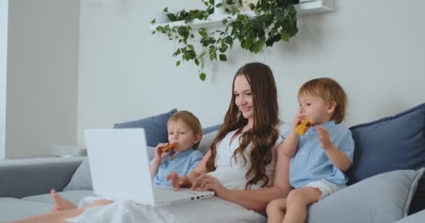 Fiatal gyönyörű anya és két fiatal gyerek ül a kanapén, látszó-on-a laptop képernyő, és hogy az online vásárlást — Stock videók