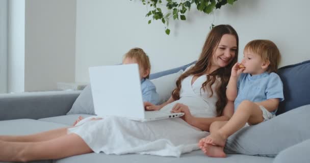 Beautiful young mom and two little kids boys are looking at the laptop screen family photos. And they do online shopping. Call grandmother through video chat. Children show their fingers on the laptop — Stock Video