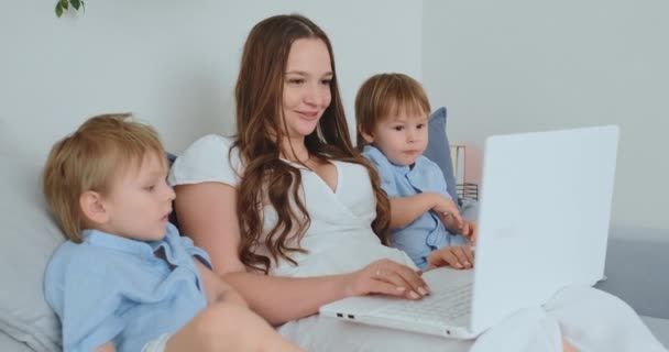 Hermosa mamá joven y dos niños pequeños niños están mirando las fotos de la familia de la pantalla del ordenador portátil. Y hacen compras en línea. Llama a la abuela a través de chat de vídeo. Los niños muestran sus dedos en el portátil — Vídeos de Stock