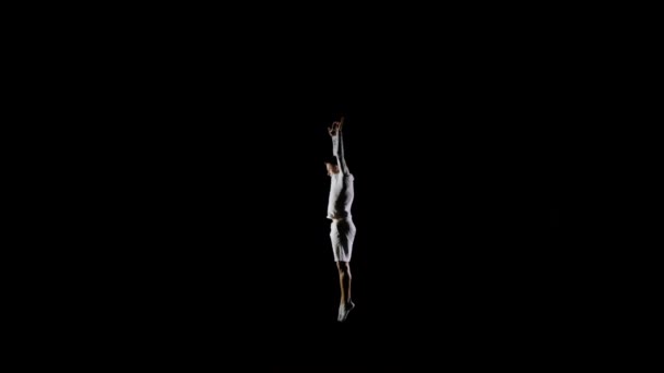Атлетик в белой одежде совершает трюки, прыгая на батуте на черном монофоническом фоне. — стоковое видео