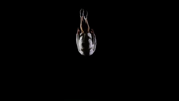 Beau gymnaste masculin en vêtements blancs sur un fond noir sautant sur un trampoline au ralenti effectuant des flips et des vis . — Video