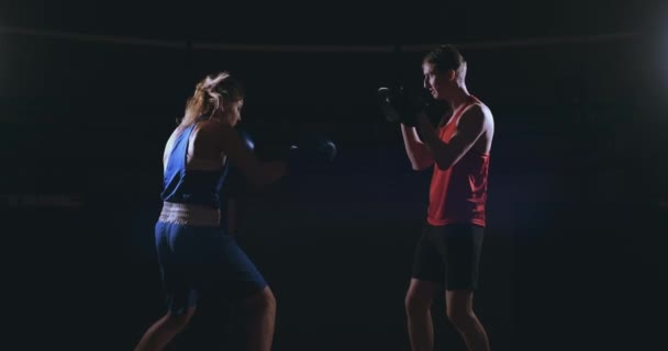 Schöne brünette Boxerin, die in einem dunklen Raum mit einem Trainer Schläge auf die Pfoten trainiert — Stockvideo