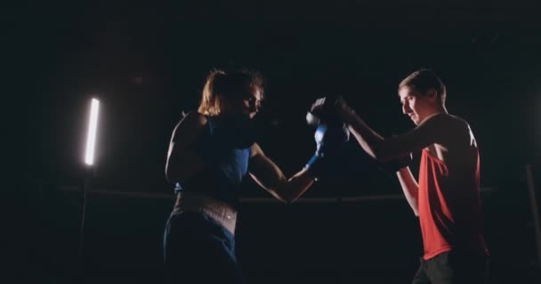 Bonito boxeador morena trabalhando golpes nas patas com um treinador em um quarto escuro — Vídeo de Stock