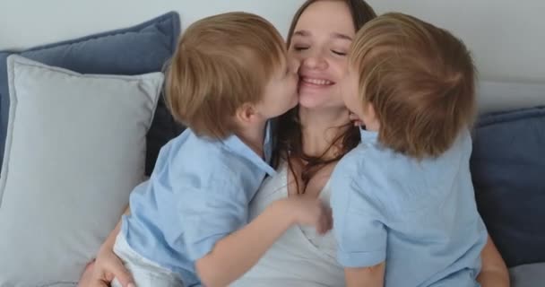 Mamá está sentada en el sofá con un portátil. Dos niños niños cantan y besan a su madre — Vídeos de Stock