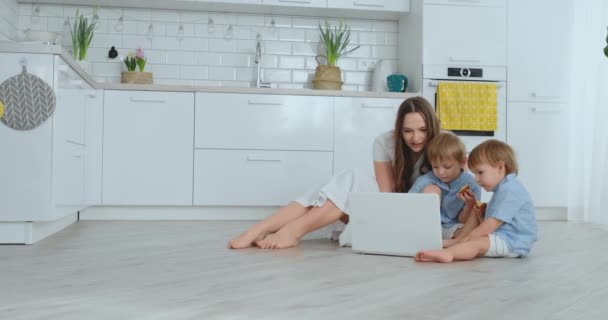 Nuori kaunis äiti istuu lattialla olohuoneessa lasten kanssa ja katselee kannettavan tietokoneen näyttöä — kuvapankkivideo