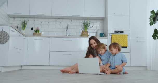 Современная квартира мама с двумя сыновьями, сидящими на полу в гостиной, смотрят на экран ноутбука — стоковое видео