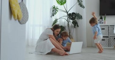 Bir dizüstü bilgisayar ile parlak bir iç modern bir dairede katta oturan iki genç çocuklu anne. Dizüstü ekran ve aile fotoğrafları seyretmek. Çocuklar için online alışveriş yapmak.