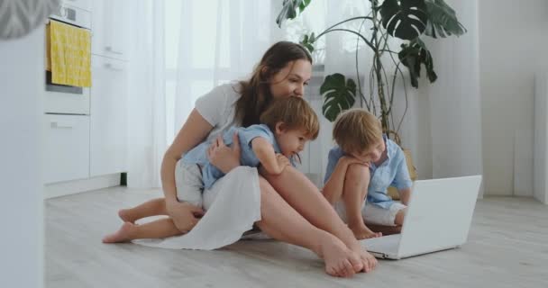 Moderno apartamento de mamá y dos hijos sentados en el suelo en la sala de estar mirar a la pantalla del ordenador portátil — Vídeos de Stock