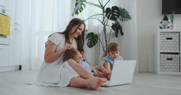 Hermosa familia joven moderna tumbada en el suelo en casa y haciendo algo en el ordenador portátil — Vídeos de Stock