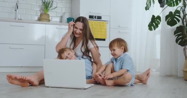 Mamma con due bambini piccoli seduti sul pavimento in un appartamento moderno in un interno luminoso con un computer portatile. Guarda lo schermo del portatile e guarda le foto di famiglia. Effettuare acquisti online per bambini . — Video Stock