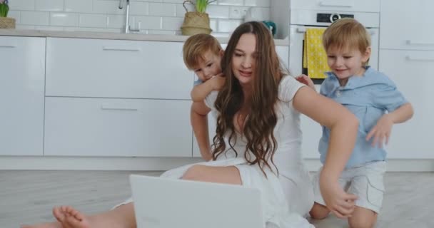 現代の技術。モダンなアパートメントの愛するママとリビング ルームの床の上に座って 2 人の小さい息子は、ノート パソコンの画面を見てください。お母さんと子供たちがラップトップでプレーします。. — ストック動画