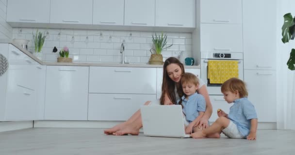 Moderno apartamento amante de mamá y dos hijos pequeños sentados en el suelo en la sala de estar mirar a la pantalla del ordenador portátil. Los niños con mamá juegan en una computadora portátil — Vídeos de Stock