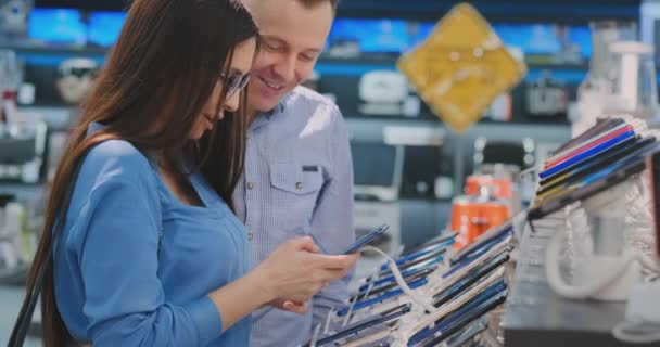 Um casal que está perto de vitrine com smartphones na moderna loja de eletrônicos e escolher um novo smartphone. Compre um telefone celular — Vídeo de Stock