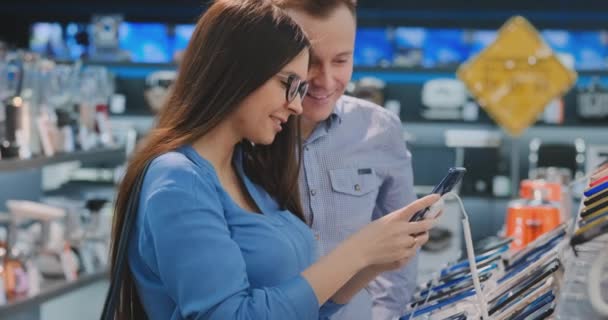 Hermosa pareja joven. Hombre y mujer tienen en las manos un teléfono inteligente de pie cerca de un escaparate con teléfonos inteligentes en la tienda de electrodomésticos elegir el mejor teléfono inteligente. Un regalo para su esposa — Vídeo de stock