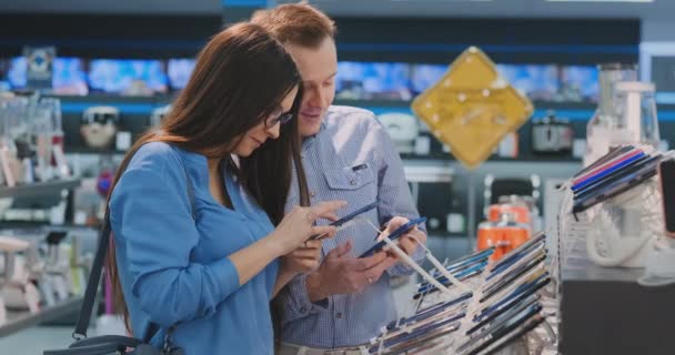 Un hombre y una mujer jóvenes tienen en sus manos dos teléfonos inteligentes de pie alrededor de un escaparate con teléfonos inteligentes en una tienda de electrónica elegir entre dos de los mejores. Comparación de dos dispositivos. Gran selección de — Vídeo de stock