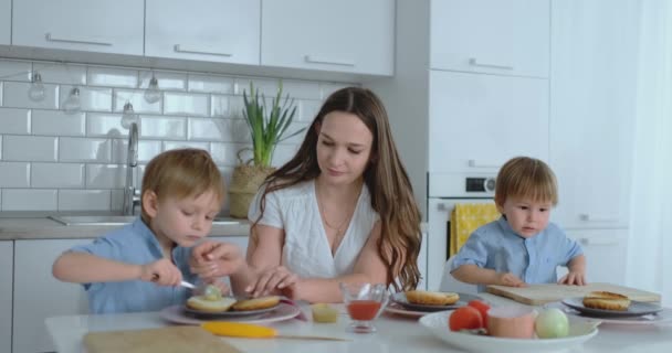 Мама с двумя детьми на кухне за столом готовит бургер к обеду — стоковое видео