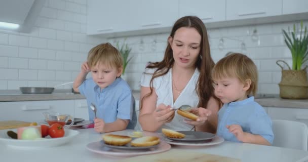 Два маленьких брата с мамой смешат бургеры и улыбаются вместе. Счастье семьи на кухне — стоковое видео