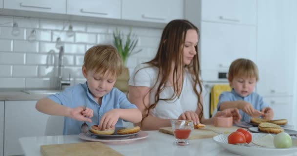 Dois irmãozinhos com sua mãe em um vestido estão cozinhando juntos em uma cozinha branca hambúrgueres rir e sorrir. Família felicidade na cozinha — Vídeo de Stock