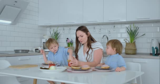 Dos niños pequeños ayudan a mamá en la cocina a preparar hamburguesas rebanando verduras, queso y salchichas — Vídeo de stock