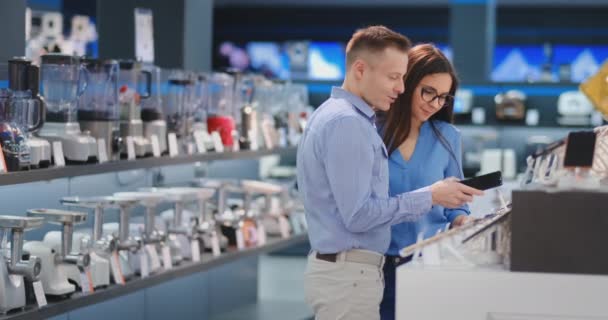 Jovem casal bonito um homem de camisa e uma mulher de óculos segurando um smartphone em pé em torno de uma vitrine com smartphones em uma loja de eletrônicos verificando a tela sensível ao toque da tela — Vídeo de Stock