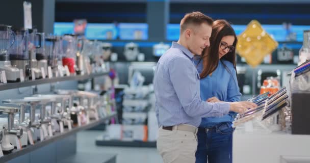 Счастливая семейная пара мужчина и женщина стоят за стойкой с мобильными телефонами в повседневной одежде, выбирая новый смартфон в современном магазине электроники — стоковое видео