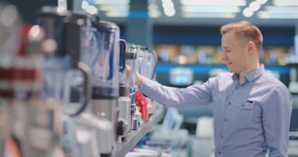 Jovem homem bonito na loja de eletrodomésticos escolhe um liquidificador para sua cozinha olhando e segurando vários modelos em suas mãos — Vídeo de Stock