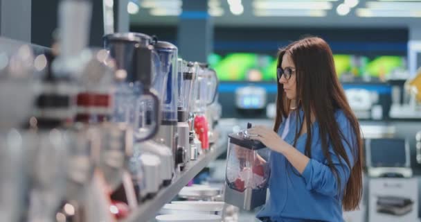 Giovane bella donna nel negozio di elettrodomestici sta scegliendo per la sua cucina un frullatore cercando e tenendo in mano vari modelli . — Video Stock