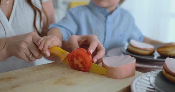 Młoda matka z małym dzieckiem razem wyciąć z nożem pomidor do domowej roboty Burger. Zdrowe jedzenie gotować razem — Wideo stockowe