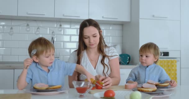 Una familia feliz es una joven y hermosa madre en un vestido blanco con dos hijos en camisas azules preparando una cocina blanca juntos rebanando verduras y creando berger saludable para los niños . — Vídeo de stock