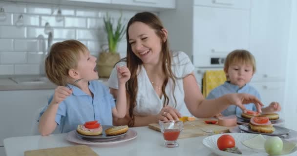 Мама с двумя детьми на кухне за столом готовит бургер к обеду — стоковое видео