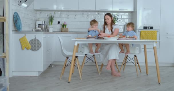 Dois meninos pequenos crianças ajudar a mãe na cozinha preparar hambúrgueres fatiar legumes queijo e salsichas — Vídeo de Stock