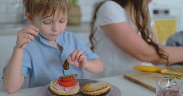 En lycklig familj är en ung vacker mor i en vit klänning med två söner i blå skjortor förbereder ett vitt kök tillsammans skivning grönsaker och skapa hälsosamma Berger för barn. — Stockvideo