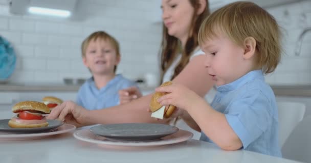 Baby pojke sitter i köket med sin mor och bror äter en hamburgare och leende. Hälsosam mat, hem hamburgare — Stockvideo