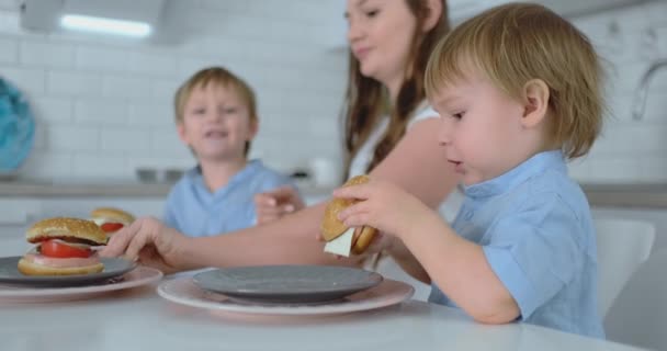 Baby Boy ül a konyhában az anyja és testvére eszik egy hamburgert és mosolyogva. Egészséges táplálkozás, házi hamburgerek — Stock videók