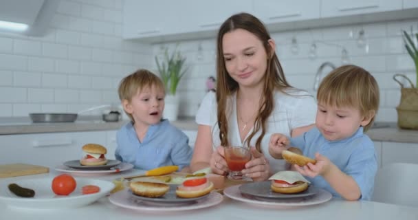 Young mother with two young sons in the kitchen at the table preparing burger for lunch — Stock Video