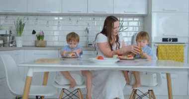 Mutlu bir aile, beyaz bir elbise, beyaz bir mutfak birlikte sebze Dilimleme ve çocuklar için sağlıklı Berger yaratma hazırlamak mavi gömlek iki oğlu ile genç bir güzel anne.