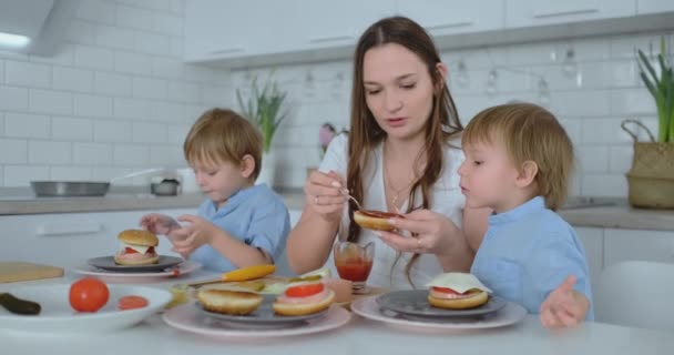 Hamburger ile beyaz ışık Kunası yemek iki çocuk oğulları ile güzel genç anne — Stok video