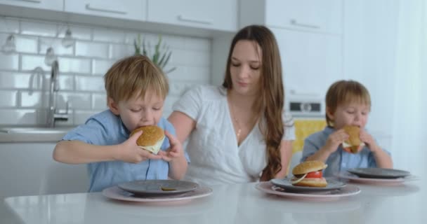 Twee kinderen in blauwe shirts eten hamburgers in de lichte keuken — Stockvideo