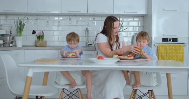 Μια ευτυχισμένη οικογένεια είναι μια νεαρή όμορφη μητέρα με ένα λευκό φόρεμα με δύο γιους με μπλε πουκάμισα που ετοιμάζουν μια λευκή κουζίνα μαζί κόβοντας λαχανικά και δημιουργώντας υγιεινό μπέργκερ για παιδιά. — Αρχείο Βίντεο
