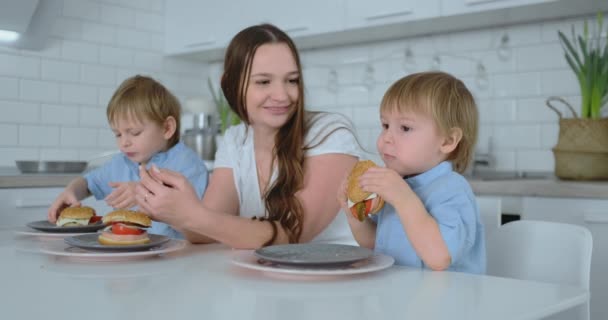 Niño sentado en la cocina con su madre y su hermano comiendo una hamburguesa y sonriendo. Comida saludable, hamburguesas caseras — Vídeo de stock