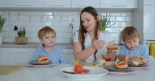 Young beautiful mom in a white dress with two children are smiling and eating fresh burgers in their kitchen — Stock Video