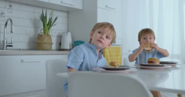 Una joven y hermosa madre en un vestido blanco con dos hijos sonríe y come hamburguesas frescas en su cocina. Familia feliz comida casera alimentos saludables — Vídeos de Stock