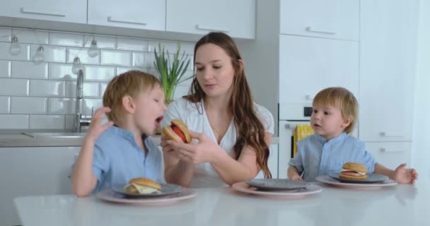 Una joven y hermosa madre en un vestido blanco con dos hijos sonríe y come hamburguesas frescas en su cocina. Familia feliz comida casera alimentos saludables — Vídeo de stock