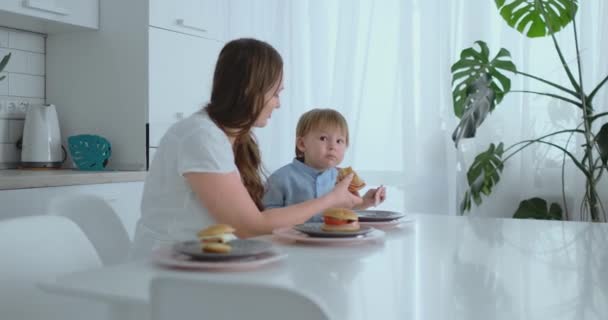 Mamma hjelper sønnen sin med å spise en burger på kjøkkenet. – stockvideo